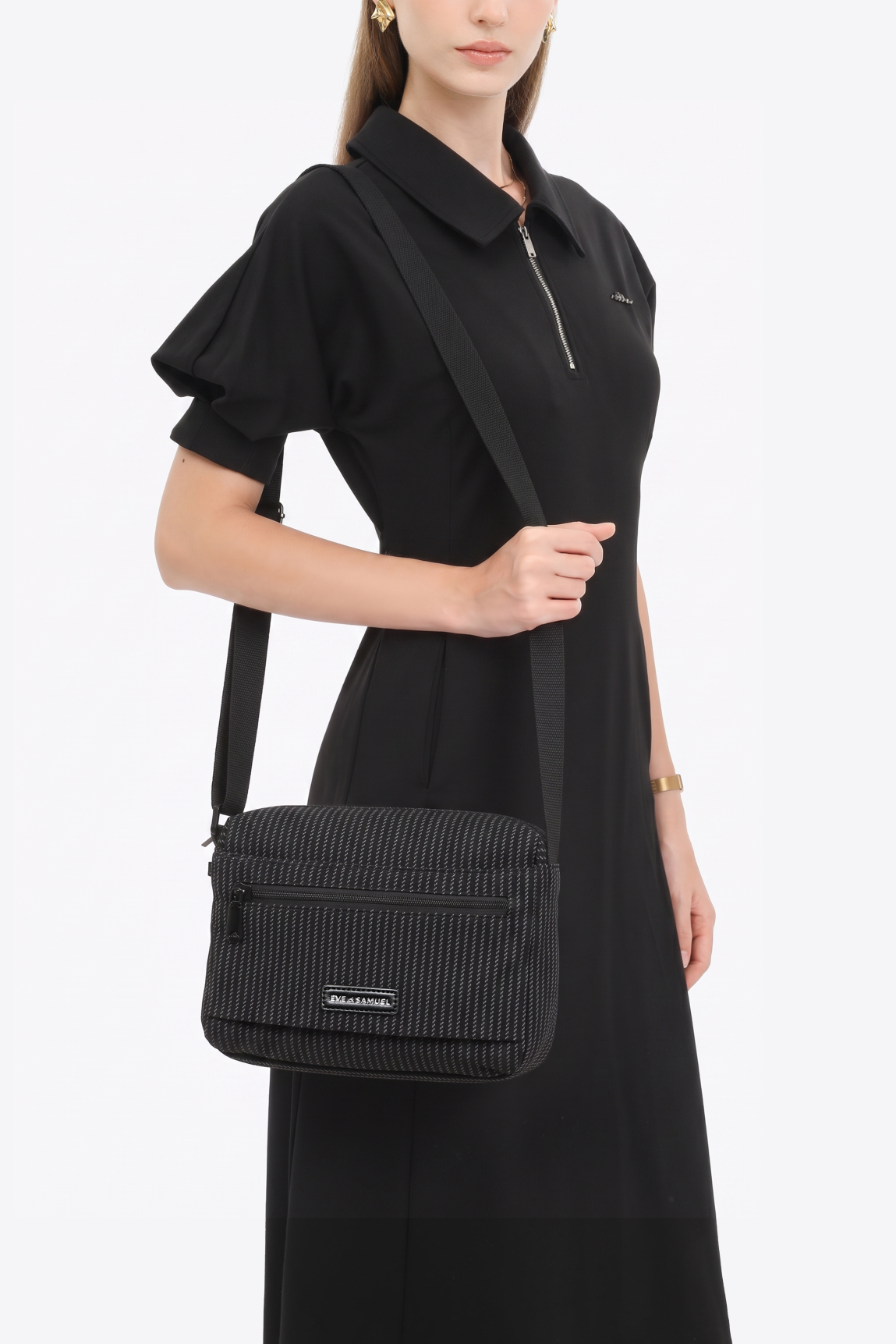 A woman modeling a black short-sleeve polo dress with a collar and front zipper, carrying a black ribbed shoulder bag with a thin strap against a plain light gray studio background.
